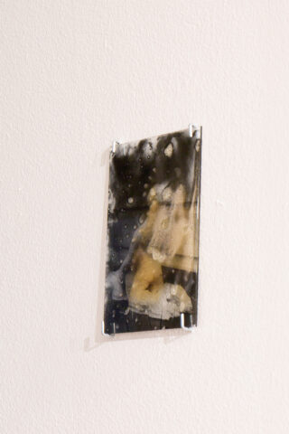 Ein kleines rechteckiges abstraktes Kunstwerk mit dunklen, wolkigen Mustern mit beigen und weißen Flecken ist mit durchsichtigen Eckhalterungen an einer weißen Wand angebracht.