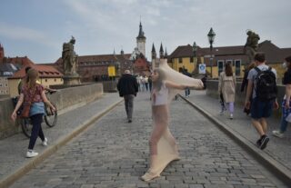 Eine in einen durchsichtigen Stoff gehüllte Person steht mit ausgestreckten Armen auf einer Kopfsteinpflasterbrücke zwischen Fußgängern in einer historischen europäischen Stadt.