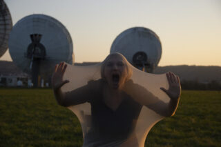 Eine Person spannt schreiend ein durchsichtiges Laken über Gesicht und Körper, im Hintergrund sind bei Sonnenuntergang große Satellitenschüsseln zu sehen.
