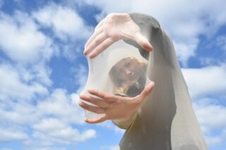 Eine Person spannt sich einen durchsichtigen Stoff über Gesicht und Hände und steht im Freien vor einem blauen Himmel mit vereinzelten Wolken im Hintergrund.