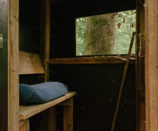 Das Innere einer Holzhütte mit einem blauen Kissen auf einer Bank, einer großen Fensteröffnung und Blick auf die Bäume draußen.