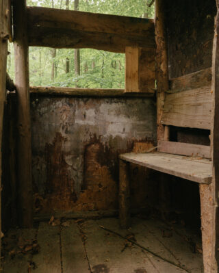 Ein kleiner, verwitterter Holzunterstand mit abblätternden Wänden und einer einfachen Bank, umgeben von dichtem grünen Wald, der durch offene Fenster sichtbar ist.