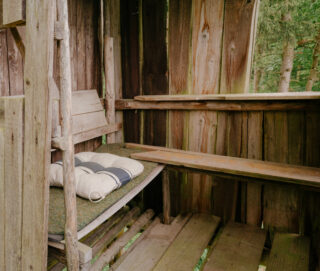 Innenansicht eines Baumhauses aus Holz mit zwei Bänken, eine davon mit einem Kissen und einer Decke bedeckt, umgeben von rauen Holzwänden und mit Blick ins Grüne draußen.