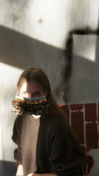 Eine Person mit langen Haaren trägt eine handgefertigte Gesichtsmaske, die mit zahlreichen Holzstäben verziert ist, und sitzt im Sonnenlicht vor einem Hintergrund mit rot und schwarz aufgemalten Formen.