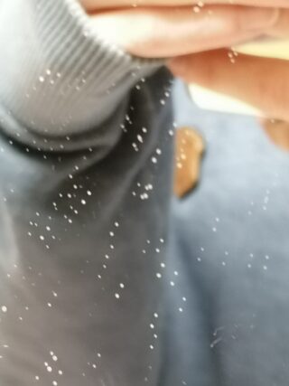 Nahaufnahme einer Person in einem grauen Pullover, die in einem Spiegel ein Foto macht, auf dessen Oberfläche sich sichtbare weiße Flecken oder Staub befinden.