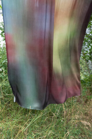 Ein halbtransparenter Stoff mit gemischten Rot-, Grün- und Brauntönen hängt im Freien über hohem Gras und Blattpflanzen.