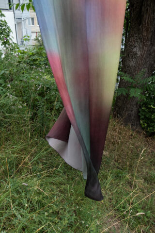 Ein mehrfarbiger Stoff mit einem Farbverlaufsmuster hängt im Freien, teilweise über Gras und Pflanzen in der Nähe eines Baumstamms drapiert.