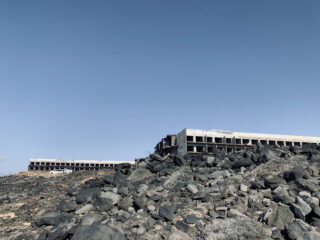 Zwei große, unfertige Betongebäude stehen auf einem felsigen, kargen Hügel unter einem klaren blauen Himmel.