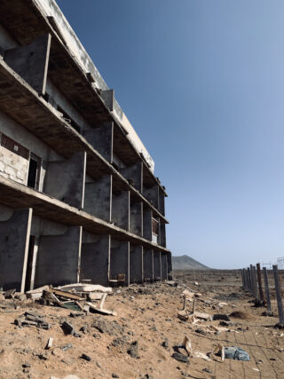 Ein teilweise fertiggestelltes, verlassenes Gebäude steht in einer kargen, felsigen Gegend, die mit Trümmern übersät ist, unter einem klaren blauen Himmel.