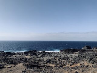 Felsige Küste mit dunklem Vulkangestein im Vordergrund, Wellen, die ans Ufer schlagen, und ein klarer blauer Himmel über einem tiefblauen Ozean.