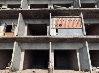 Ein teilweise fertiggestelltes, verlassenes Betongebäude mit fehlenden Wänden, offenen Fenstern und zwei leeren Stühlen davor.