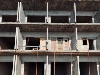 Unfertige Betonfassade eines Gebäudes mit sichtbaren Balken, Säulen und offenen Fensterflächen; einige Fenster sind teilweise mit Holz und Kunststoff abgedeckt.