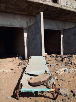 Ein beschädigtes Feldbett mit zerrissenem Stoff und einer kaputten Trommel steht auf Trümmern vor einem teilweise errichteten Betongebäude.