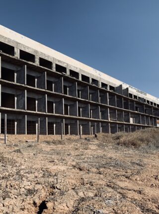 Ein großes, unfertiges Betongebäude steht auf trockenem, kargem Boden unter einem klaren, blauen Himmel.