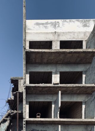 Ein teilweise fertiggestelltes oder verlassenes Betongebäude mit leeren Fensteröffnungen und einem einzelnen Stuhl auf einer Etage vor einem klaren blauen Himmel.