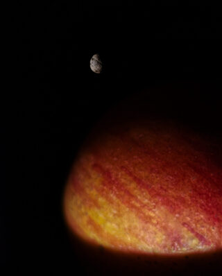 Ein kleiner, mit Kratern übersäter Mond schwebt in der Dunkelheit über einem großen, rot-orangen, planetenähnlichen Objekt mit feinen Streifen auf der Oberfläche.