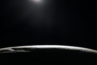 Ein gekrümmtes Objekt wird vor einem dunklen Hintergrund schwach von oben beleuchtet, mit einer hellen Lichtquelle nahe der oberen Bildkante.