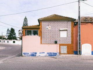 An einer Straßenecke steht ein kleines zweistöckiges Haus mit gemusterten Fliesen und pfirsichfarbenen Wänden, mit geschlossenem Fenster und einer Satellitenschüssel an der Seite.
