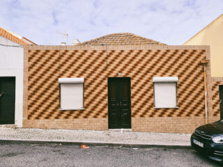 Ein einstöckiges Haus mit einer gemusterten Ziegelfassade, zwei Fenstern mit weißen Fensterläden und einer dunklen Tür in der Mitte; rechts ist teilweise ein schwarzes Auto geparkt.