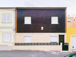 Ein zweistöckiges Haus mit einer dunkel gefliesten oberen Fassade und einer hellen unteren Fassade mit geschlossenen weißen Fensterläden und einem schwarzen Zaun entlang der Vorderseite.