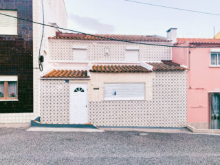 Ein kleines Haus mit gemusterter Ziegelfassade, weißer Tür und Fensterläden steht in einer ruhigen Straße zwischen dunkelbraunen und rosafarbenen Nachbargebäuden.