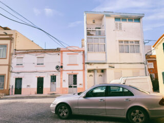 Ein silbernes Auto parkt unter klarem Himmel auf einer Straße vor pastellfarbenen Wohnhäusern mit geschlossenen Fensterläden und Balkonen.