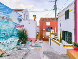 Eine schmale, farbenfrohe Straße mit leuchtenden Wandmalereien, zwischen den Gebäuden hängender Wäsche und Topfpflanzen am Wegesrand in einem mediterranen Dorf.
