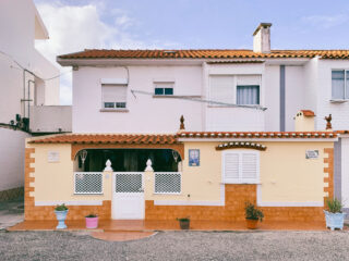Ein zweistöckiges Haus mit weißen oberen und gelben unteren Wänden, orangefarbenen Dachziegeln, weißen Fensterläden, Topfpflanzen und einem kleinen Eingangstor davor.