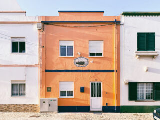 Ein schmales orangefarbenes Haus mit weißen Verzierungen, zwei Fensterläden, einer weißen Tür und einem dekorativen Fliesenwandbild an der Fassade, zwischen zwei weißen Häusern gelegen.