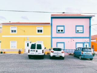 An einem klaren Tag parken zwei Autos und ein Lieferwagen vor einem gelben und einem rosa Haus mit blauen Verzierungen.