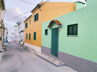 Schmale Straße gesäumt von farbenfrohen Gebäuden, darunter gelbe und grüne Fassaden, mit geschlossenen Fensterläden und einem Perlenvorhang über einem Eingang.