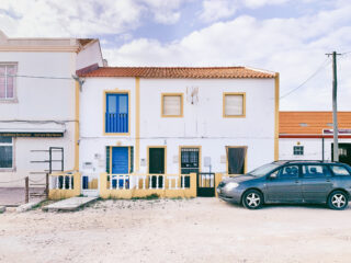 Ein zweistöckiges weißes Haus mit einem roten Ziegeldach, einer blauen Haustür, einem kleinen Balkon und einem geparkten grauen Auto davor an einem bewölkten Tag.