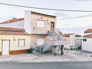 Zweistöckiges Haus mit gemusterten Fliesen und einer Außentreppe, gelegen in einer ruhigen Straße mit Nachbarhäusern und sichtbaren Versorgungsleitungen darüber.