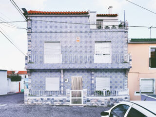 Ein zweistöckiges Haus mit einer blau-weißen Ziegelfassade, geschlossenen weißen Fensterläden, einem roten Ziegeldach und einem weißen Tor, vor dem ein weißes Auto geparkt ist.