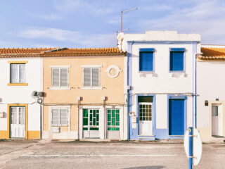 Zwei benachbarte Häuser, eines beige mit weißen Fensterläden und eines weiß mit blauen Fensterläden, stehen nebeneinander in einer sonnigen Straße unter klarem Himmel.