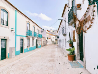 Eine schmale, leere Kopfsteinpflasterstraße, gesäumt von weißen Gebäuden, blauen Verzierungen, Balkonen, Topfpflanzen und einem dekorativen Stoffdach auf der rechten Seite.