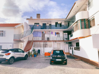 Zwei Autos parken vor einem weißen, mehrstöckigen Wohnhaus mit Balkonen, Außentreppen und Wäsche, die auf Wäscheleinen hängt.