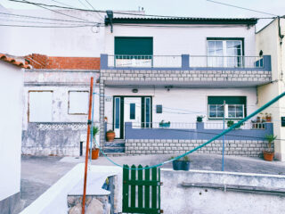 Zweistöckiges weißes Haus mit Balkon, grünen Fensterläden und Topfpflanzen am Eingang; grünes Tor und Wäscheleine im Vordergrund.