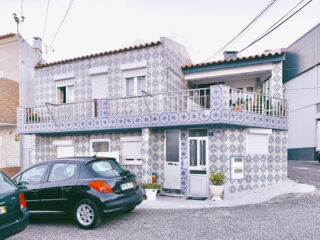 Ein zweistöckiges Haus mit einer gemusterten blau-weißen Ziegelfassade, einem Balkon und zwei davor geparkten Autos an einer gepflasterten Straße.