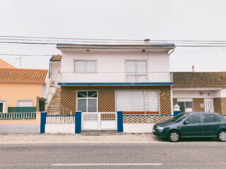 Zweistöckiges Haus mit braun-weiß karierter Fassade und blauen Verzierungen. Davor parkt ein schwarzes Auto auf einer gepflasterten Straße. Angrenzende gelb-weiße Gebäude sind sichtbar.