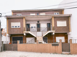 Zweistöckiges Haus mit eingezäuntem Vorgarten, brauner Außenfassade, drei Treppen, die zum Haupteingang führen, und mehreren Fenstern mit Fensterläden.