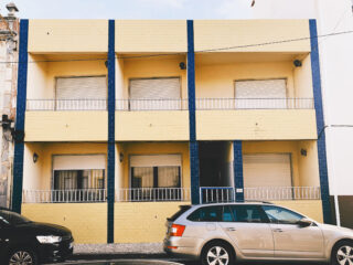 Ein zweistöckiges gelbes Gebäude mit blauen vertikalen Akzenten, geschlossenen Fensterläden an allen Fenstern und zwei davor auf der Straße geparkten Autos.