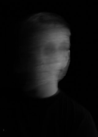 Ein unscharfes Graustufenfoto des Gesichts einer Person vor einem dunklen Hintergrund, das aufgrund der Bewegungsunschärfe ein geisterhaftes, undeutliches Aussehen erzeugt.