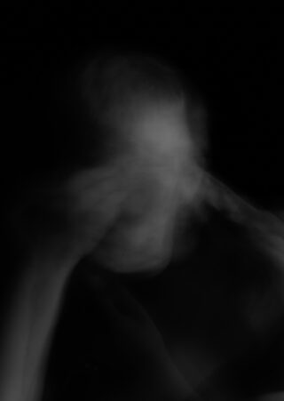 Auf einem unscharfen Schwarzweißfoto ist eine Person zu sehen, die ihre Hand vors Gesicht hält. Durch die Bewegung entsteht vor einem dunklen Hintergrund ein geisterhafter, undeutlicher Effekt.