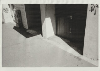 Ein Schatten fällt über einen Gehweg vor einer geschlossenen dunklen Tür und einer weißen Wand. An der Wand und einem Versorgungskasten sind Graffiti zu sehen.