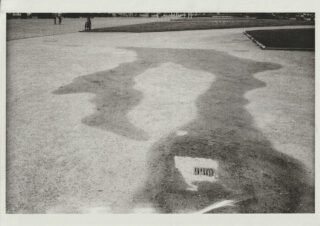 Ein großer, unregelmäßiger Schatten wird auf einen Sandweg in einem Park geworfen. Im unteren Vordergrund ist teilweise ein Buch oder eine Zeitschrift zu sehen.