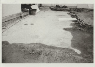 Eine Baustelle mit verstreuten Ziegeln, Schutt und Baumaterialien auf einem teilweise befestigten Untergrund neben einer gefliesten Fläche.