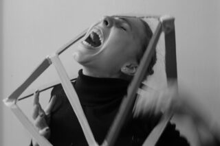 Eine Person im Rollkragenpullover schreit mit offenem Mund und hält einen geometrischen Holzrahmen um den Kopf. Das Foto ist schwarz-weiß.