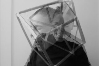 Ein Schwarzweißfoto mit Doppelbelichtung zeigt eine schreiende Person in einer geometrischen Pappstruktur, wobei ihr Gesicht an zwei sich überlappenden Positionen sichtbar ist.
