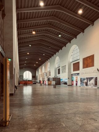 Große, größtenteils leere Bahnhofshalle mit hohen Bogenfenstern, einer Reihe von Werbeanzeigen entlang einer Wand und einem gefliesten Boden unter einer gerippten Decke.